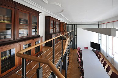 Sehr hoher Bibliotheksraum im modernisierten Altbau mit Fischgrätparkett. Auf einer durch schwarze gusseiserene Säulen gestützten, schmalen Galerie stehen historische Holzvitrinen mit ledergebundenen Büchern darin. Auf der unteren Ebene nimmt ein grauer Besprechungstisch die gesamte Länge des Raums ein.