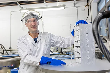 Genlabor im Zentrum für Seltene Immunologische Erkrankungen Mann im weißen Laborkittel, transparentem Visir und blauen Schutzhandschuhen hebt ein angefrorenes Gestell mit Schubladen aus einem runden Gefrierbehälter mit flüssigem Stickstoff, aus dem Dampf aufsteigt.