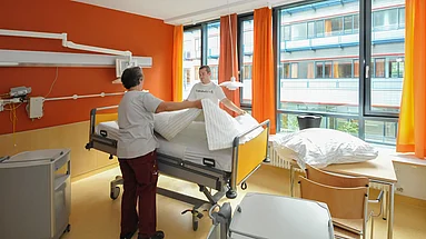 2 Mitarbeitende beim Bettenwechsel in einem in warmen orange gehaltenen Patientenzimmer mit großer Fensterfront.