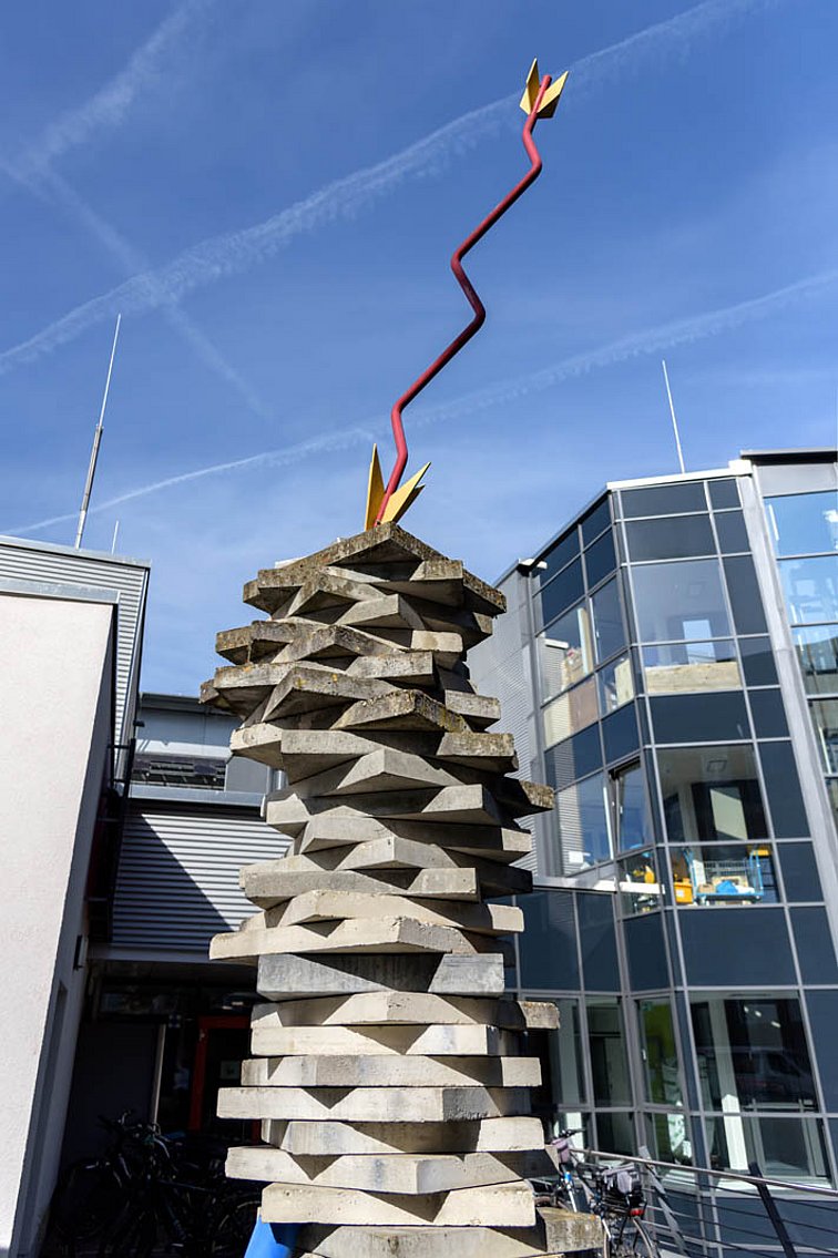 Ein übergroßer rot-gelber Pfeil steht mit der Spitze nach unten auf einem Haufen aufeinander gestapelter Steinplatten.