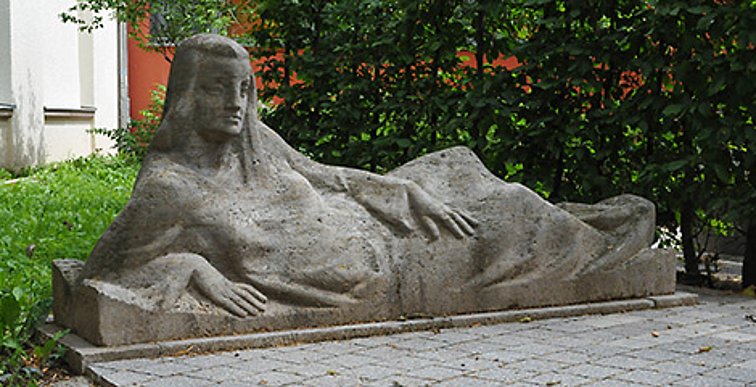 Steinfigur einer entspannt seitlich auf den Arm gestützt liegenden Frau.