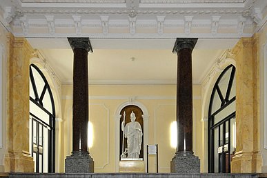 Historischer, gelb gestrichener Eingangsbereich im Pathologischen Institut mit zwei dunklen Säulen und einer weißen Statue mit Toga, Helm und Speer auf einem Sockel.