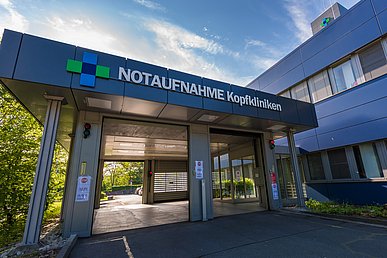 Zweiseitig befahrbare Halle mit der Beschriftung Notaufnahme Kopfkliniken. Nach rechts aus der Halle erfolgt der direkte Zugang zum Klinikgebäude, dessen Fassade mit dunkelblauen Metallelementen verkleidet ist.