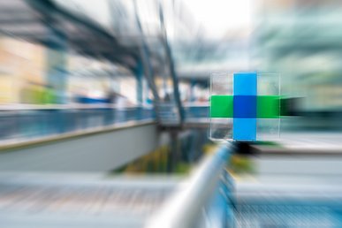 Das kreuzförmige Logo des Uniklinikums Erlangen dreidimensional in einen Plexiglaswürfel gegossen. In der Mitte des Kreuzes ein dunkelblaues Quadrat, die Flügelquadrate oben und unten sind hellblau, die links und rechts grün. Unscharf im Hintergrund der Außenbereich eines modernen Klinikgebäudes.