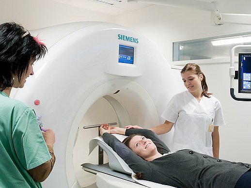 2 Mitarbeiterinnen sprechen mit einer lächelnden Patientin die auf der Liege des CT-Gerätes liegt.