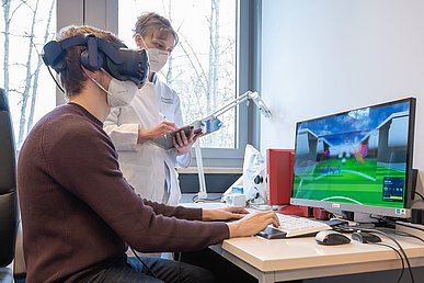 Post-COVID-Forschung in der Augenklinik Ein junger Mann mit VR-Brille sitzt vor einem Bildschirm, auf dem ein grünes Spielfeld zu sehen ist und macht auf Eingaben auf einer Tastatur. Eins stehend junge Frau in weißem Kittel beobachtet ihn und macht Notizen auf einen Klemmbrett.
