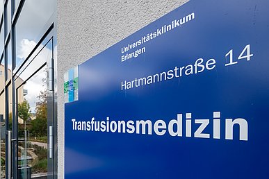 In schräger Perspektive aufgenommenes, dunkelblaues Eingangsschild mit weißer Beschriftung „Transfusionsmedizin“ und Logowürfel des Uniklinikums Erlangen aus Plexiglas vor der gläsernen Eingangstür des Gebäudes.
