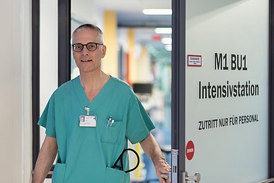 Lächelnder Mann mit Brille, grauen Haaren und grüner Funktionskleidung blickt direkt in die Kamera während er nach außen eine Türe aus Milchglas mit der Aufschrift Intensivstation öffnet. 