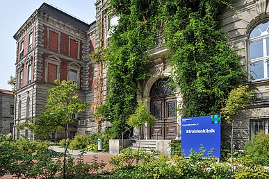 Vor der grün bewachsenen roten Klinkerfassade des historischen Gebäudes im Stil der Gründerzeit steht ein quadratisches, blaues Gebäudeschild mit dem Schriftzug Strahlenklinik.