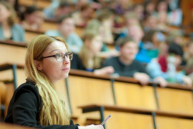Porträtaufnahme eine Studentin, die konzentriert nach vorne blickt. Im Hintergrund unscharf weitere Studenten, die in Hörsaalbänken aus hellem Holz sitzen.
