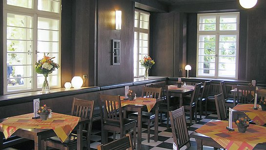 Blick in einen mit Schachbrettmuster gefliesten Raum mit dunkelbrauner Holztäfelung und gleichfarbigen Möbeln. Auf den Tischen sind orange gemusterte Decken und Blumen stehen vor den weißen Fenstern.