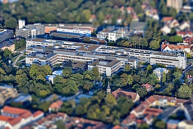 Luftaufnahme von West nach Ost. Das quer liegende graublaue Internistische Zentrum in baumbestandener Umgebung ist scharf abgebildet. Vordergrund und Hintergrund sind unscharf.