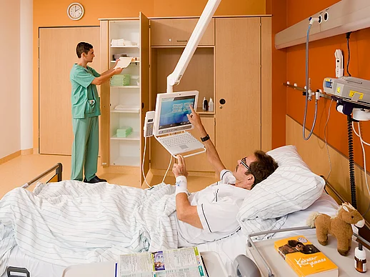 Patient liegt im Bett in einem warm-orangen Zimmer und bedient mobilen Bildschirm, während Pfleger im Hintergrund etwas aus dem Schrank holt.
