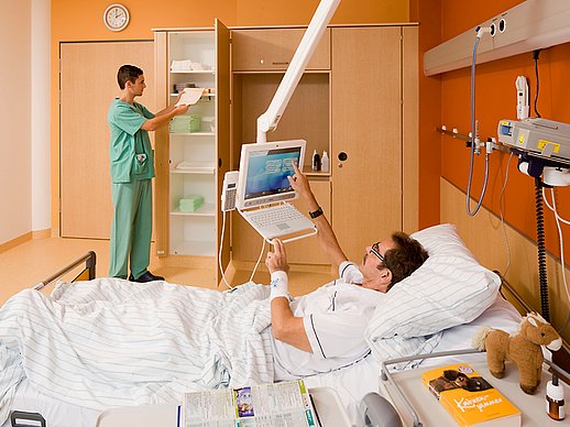 Patient liegt im Bett in einem warm-orangen Zimmer und bedient mobilen Bildschirm, während Pfleger im Hintergrund etwas aus dem Schrank holt.