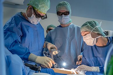 Ein Operateur mit Polarisationsbrille bewegt ein stabförmiges Gerät an dessen Spitze eine helle, weiße Lichtquelle sitzt in den Bauchraum eines Patienten. Ein Mann und eine Frau, alle in blauer und grüner Operationskleidung assistieren mit weiteren Instrumenten.
