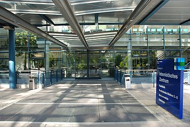 Blick vom überdachten Zugang des internistischen Zentrums auf die Glasfassade des Eingangsbereichs mit Schiebetüren. Rechts das dunkelblaue Eingangsschild mit den Bezeichnungen der darin befindlichen Einrichtungen in weißer Schrift: Hautklinik, Medizinische Kliniken 1 bis 5, Nuklearmedizin und Radiologie.
