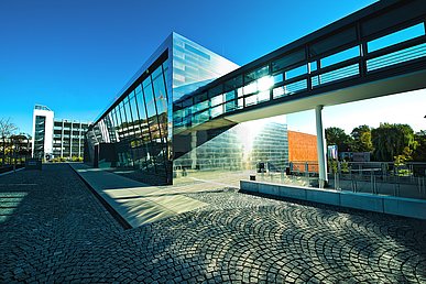 In die mit spiegelnden Metallplatten verkleidete Südfront des kubusförmigen Gebäudes der Hörsäle Medizin ragt im ersten Stock ein Verbindungsgang aus Glas und Stahl. Die Westseite ist verglast.