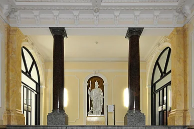 Historischer, gelb gestrichener Eingangsbereich im Pathologischen Institut mit zwei dunklen Säulen und einer weißen Statue mit Toga, Helm und Speer auf einem Sockel.