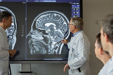 Arztkonferenz in einen Besprechungsraum: Ein Mediziner erläutert seinen rechts und links stehenden Kollegen Details zu einen Hirnscan, der auf einem großen Wandbildschirm zu sehen ist.