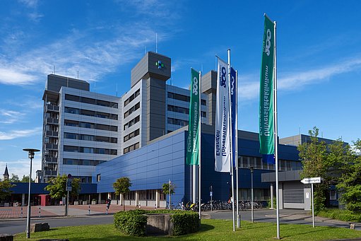Blick auf das achtstöckige, hochhausartige Gebäude der Kopfkliniken mit hell und dunkelblauer, metallverkleideter Fassade. Vorgelagert ein zweigeschossiger Gebäudeteil mit dunkelblauer Metallfassade. Im Vordergrund ein Fahnenwald mit zwei grünen einer weißen und einer blauen Fahne des Universitätsklinikums Erlangen.