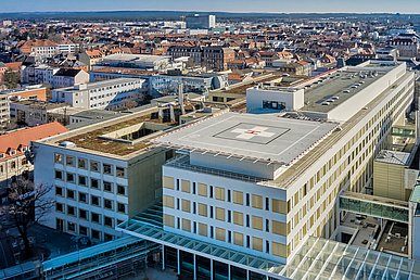 Luftaufnahme von schräg oben auf das fünfstöckige Chirurgische Zentrum mit weißer Fassade mit goldgelben Fenstereinfassungen. Im Vordergrund das auf dem Dach platzierte Quadrat des Hubschrauberlandeplatzes.