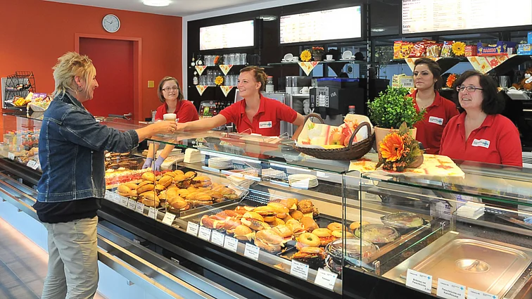 Frau bekommt von lächelnder Mitarbeiterin Kaffee übergeben. In Glastheke sind belegte Brötchen und Kuchen zu sehen. Die Wände im Hintergrund sind orange bzw. schwarz und mit Sonnenblumen dekoriert.