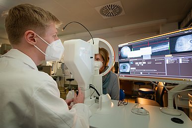 Post-COVID-Forschung in der Augenklinik Junger Mann mit weißem Kittel und weißer FFP2-Maske führt bei einer Frau, die ebenfalls Maske trägt bei gedämpften Licht eine Augenuntersuchung durch. Die Frau blickt in das kreisförmige Gerät, das Augeninnere wird auf einen Bildschirm visualisiert.