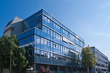 Das würfelförmige Gebäude der Zahnkliniken vor blauen Himmel von unten fotografiert. In der glänzenden Fassade spiegeln sich der blaue Himmel und weiße Wolken.