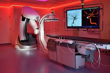 Blick auf ein Angiographie-Gerät, bestehend aus eine Liege und einem zangenförmigen Untersuchungsgerät, das über einen großen Steuerarm in verschiedenste Positionen gefahren werden kann, in einen rot ausgeleuchteten Untersuchungsraum. Auf zwei großen Monitoren sind Angiographien zu sehen.
