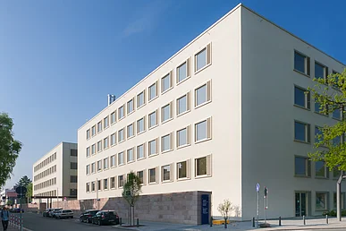 Das in zwei Würfel gegliederte fünfstöckige Gebäude des Chirurgischen Bettenhauses mit weißer Fassade und golden eloxierten Fensterrahmen aus Aluminium vor blauem Himmel. Im Vordergrund die wieder aufgebaute historische östliche Stadtmauer aus Sandsteinquadern.