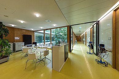 Stationsflur und Aufenthaltsbereich mit Blick auf den verglasten und begrünten Innenhof. Türen, teils auch Wände sind holzgetäfelt, der Boden ist gelb, die weiteren Wände und die Tische und Stühle sind weiß.