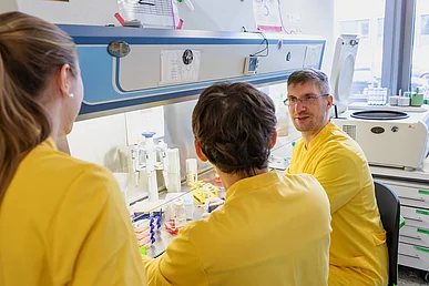Laborarbeit im Zentrum für Seltene Immunologische Erkrankungen Drei Labormitarbeiter in gelben Laborkitteln sitzen und stehen diskutierend an einer Laborbank mit verschiedenen Reagenzien.