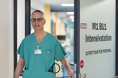 Lächelnder Mann mit Brille, grauen Haaren und grüner Funktionskleidung blickt direkt in die Kamera während er nach außen eine Türe aus Milchglas mit der Aufschrift Intensivstation öffnet. 