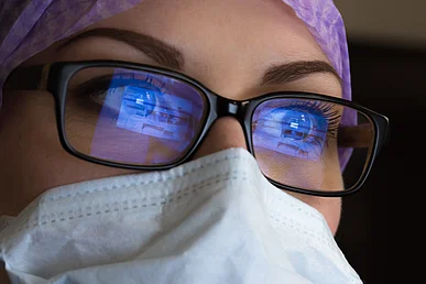 Nahaufnahme des Gesichts einer Frau mit Mundschutz und violetter OP-Haube. In ihrer Brille mit schwarzer Fassung spiegelt sich vor beiden Augen bläulich ein Computerbildschirm.