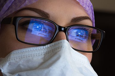 Nahaufnahme des Gesichts einer Frau mit Mundschutz und violetter OP-Haube. In ihrer Brille mit schwarzer Fassung spiegelt sich vor beiden Augen bläulich ein Computerbildschirm.