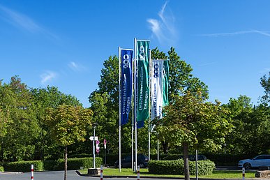 Fahnenwald mit blauen, grünen und weißen Fahnen mit der Aufschrift  Universitätsklinikum Erlangen 200 Jahre seit 1815 vor einer Grünanlage mit Bäumen im Hintergrund.