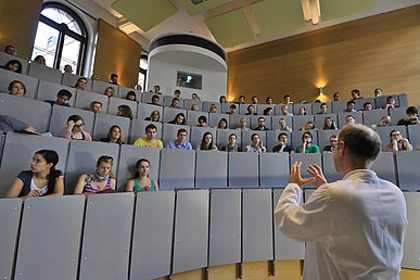 Dozent von hinten aufgenommen steht in einem Hörsaal gestikulierend vor Studierenden. Die grauen Sitzreihen sind viertelkreisförmig angeordnet.