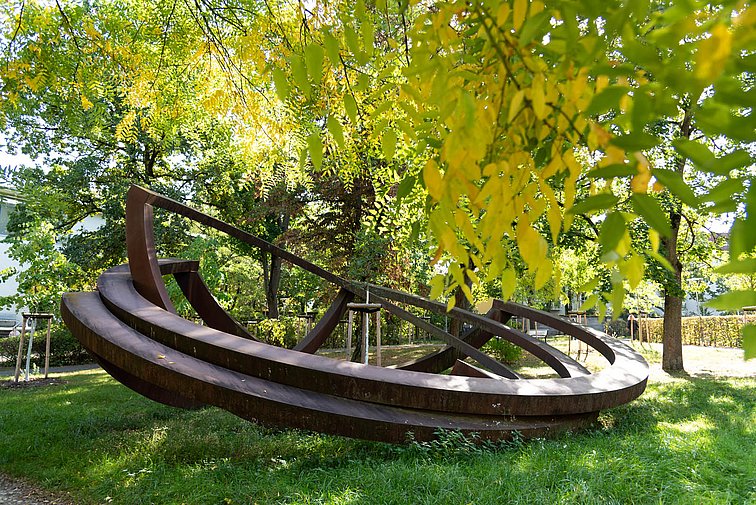 Das Kunstwerk besteht aus drei ca. 6 Meter breiten bronzefarbenen Metall-Ringen, die an einer Seite wie zusammengefaltet abknicken.