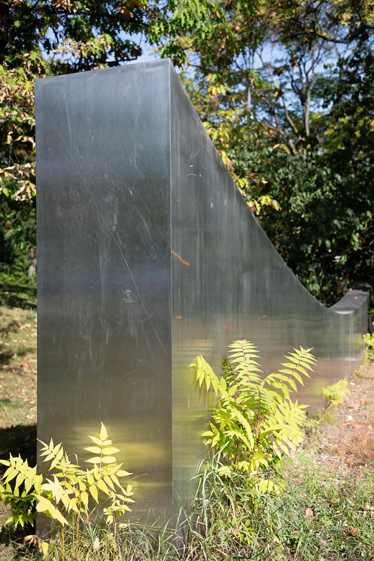 Die Skulptur gleicht einer Metallwand die in einem Bogen - mit der tiefsten Stellen in der Mitte - abgeschnitten wurde.