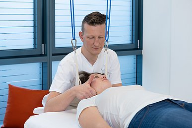 Physiotherapeut sitzt hinter einer Patientin auf einer Behandlungsliege. Der Hinterkopf der Patientin ist auf er Schlinge gelagert, der Therapeut greift an das Kiefergelenk der Patientin.