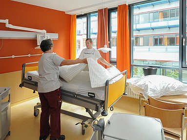 2 Mitarbeitende beim Bettenwechsel in einem in warmen orange gehaltenen Patientenzimmer mit großer Fensterfront.