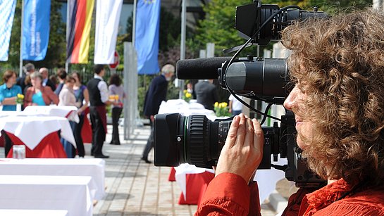 Mitarbeiterin mit professioneller Kamera samt Mikrofon filmt eine Veranstaltung.