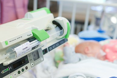 Medikamentensicherheit: Forschung in der Kinder- und Jugendklinik Links im Vordergrund eine Spritzenpumpe mit eingelegtem Medikament. Im Hintergrund unscharf ein Kleinkind im Gitterbettchen.