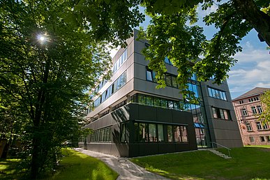 Zwischen vom Sonnenlicht durchdrungenen Bäumen Blick auf die braun eloxierte Fassade des modernen Translational Research Centers.