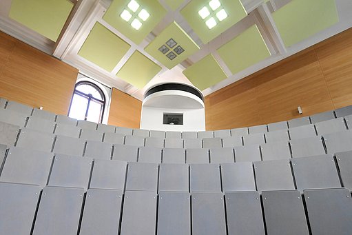 Modernisierter historische Hörsaal mit halbkreisförmigen grauen Sitzreihen, holzgetäfelten Wänden und einer lindgrünen Kassettendecke.