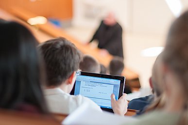 Ein Student im Hörsaal blickt auf ein Tablet auf dem ein Dokument auf sichtbar ist. Vor und hinter dem Studenten sind unscharf weitere Studierende und der Dozent zu sehen.