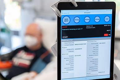 Auf einen Tablet ist der Fortschritt einer transfusionsmedizinischen Behandlung abzulesen. Ich Hintergrund ist der Patient unscharf auf einer Behandlungsliege zu sehen.