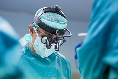 Operateur in OP-Kleidung mit Mundschutz, Haube und Mikroskop-Brille blickt nach unten auf den OP-Tisch.