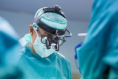 Operateur in OP-Kleidung mit Mundschutz, Haube und Mikroskop-Brille blickt nach unten auf den OP-Tisch.