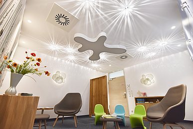 Weiß gestrichener Raum mit strahlenförmigen Lichteffekten an der Decke. Möbliert mit Holzmöbeln, bräunlichen Sesseln und Kindersesseln aus Kunststoff in Grün und Blau. 